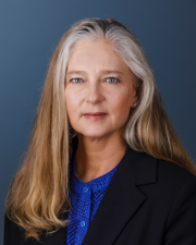 Woman with light hair in a suite jacket and blue shirt looking at the camera on a blue background